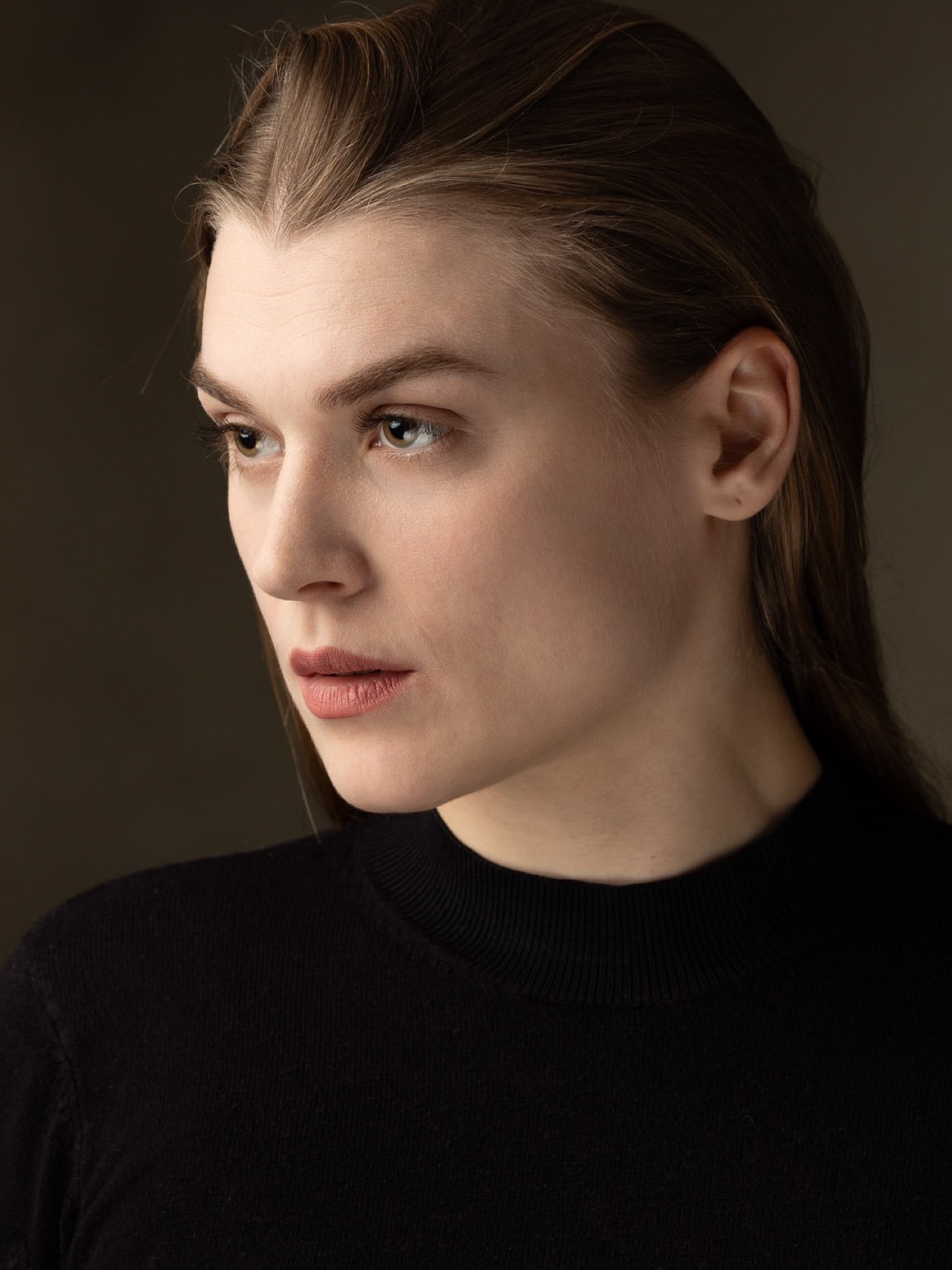 Portrait of a woman with slicked-back hair in a black mock-neck sweater, looking off camera against an olive backdrop
