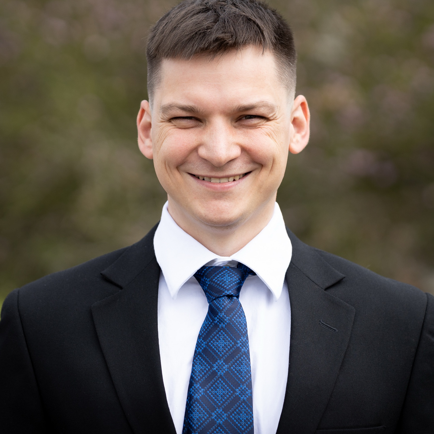 Corporate headshot of a smiling man in a dark suit and blue patterned tie, photographed outdoors