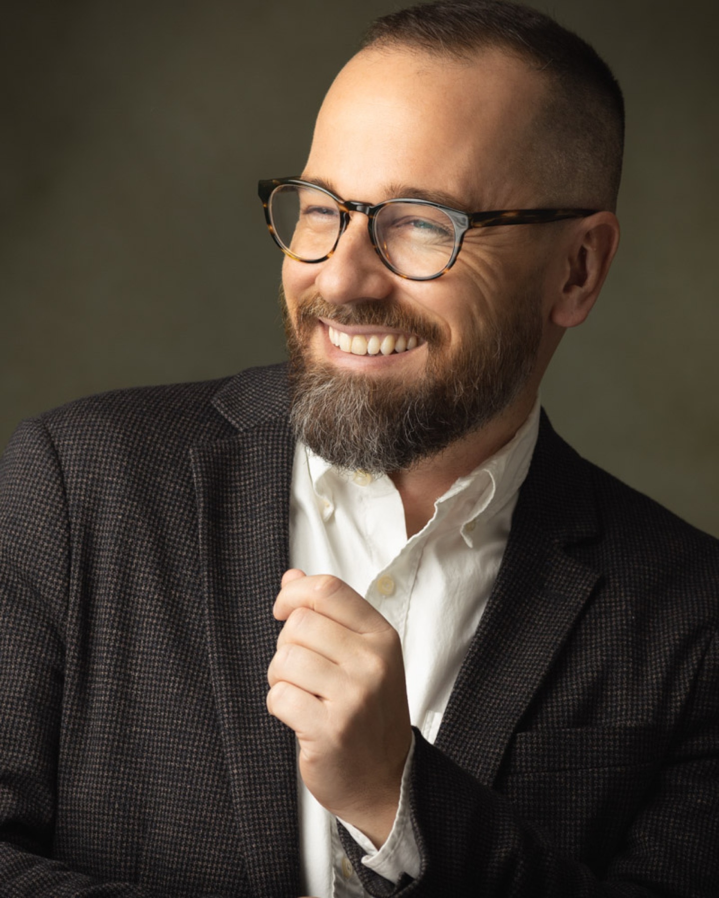 Smiling man in glasses and a tweed blazer photographed against an olive backdrop