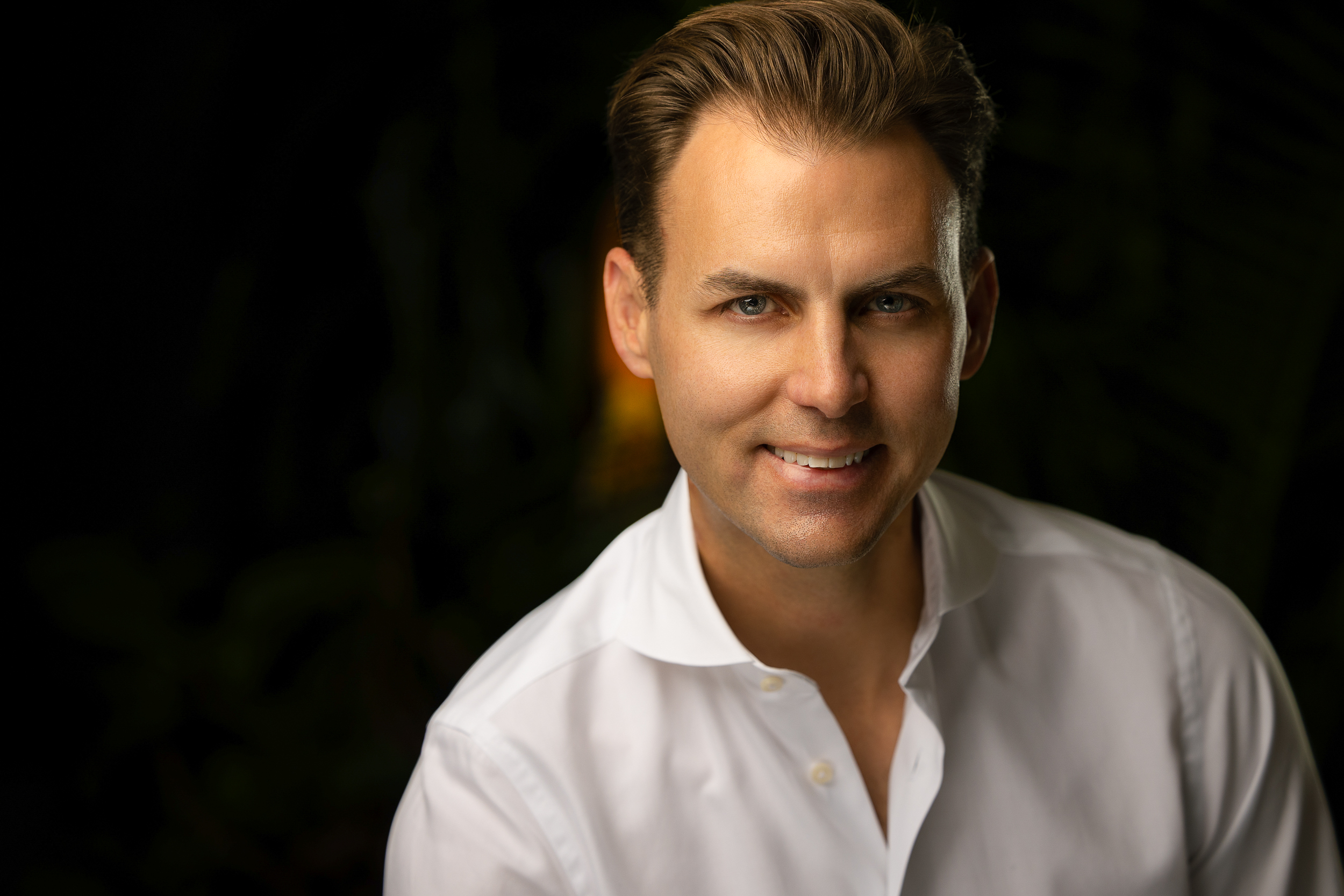 Studio portrait of a smiling man in an open-collar white shirt against a dark background