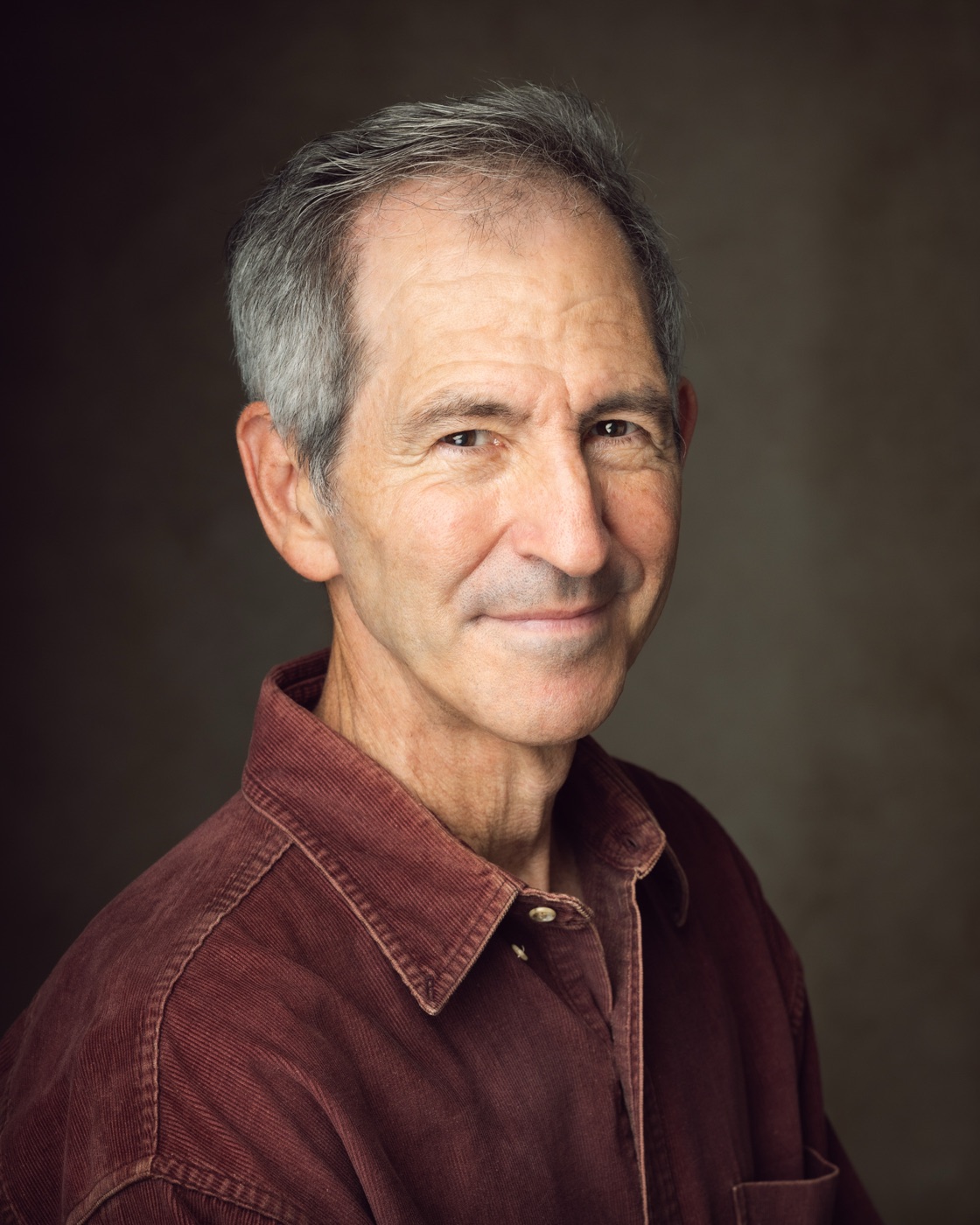 Smiling grey-haired man in a maroon corduroy shirt against a warm studio backdrop