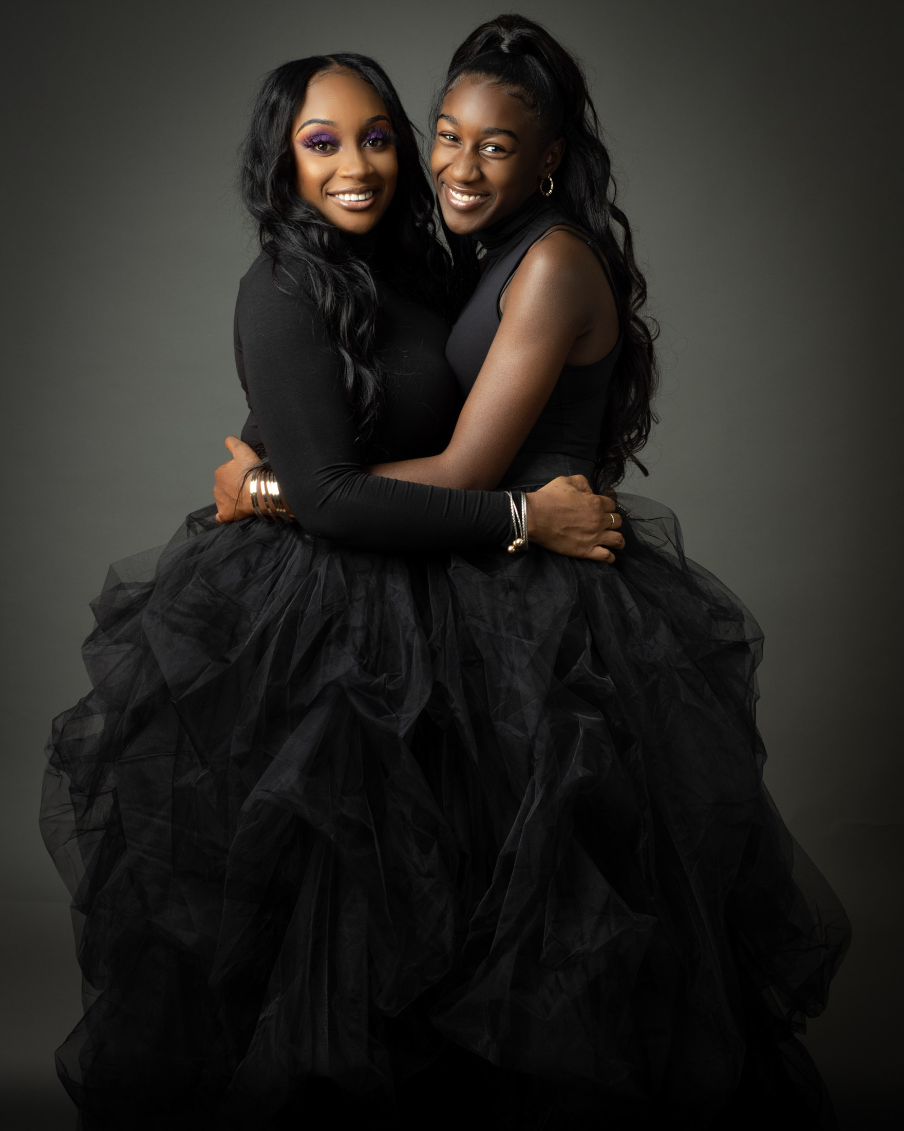 Two women in black formal dresses smiling together in a studio portrait